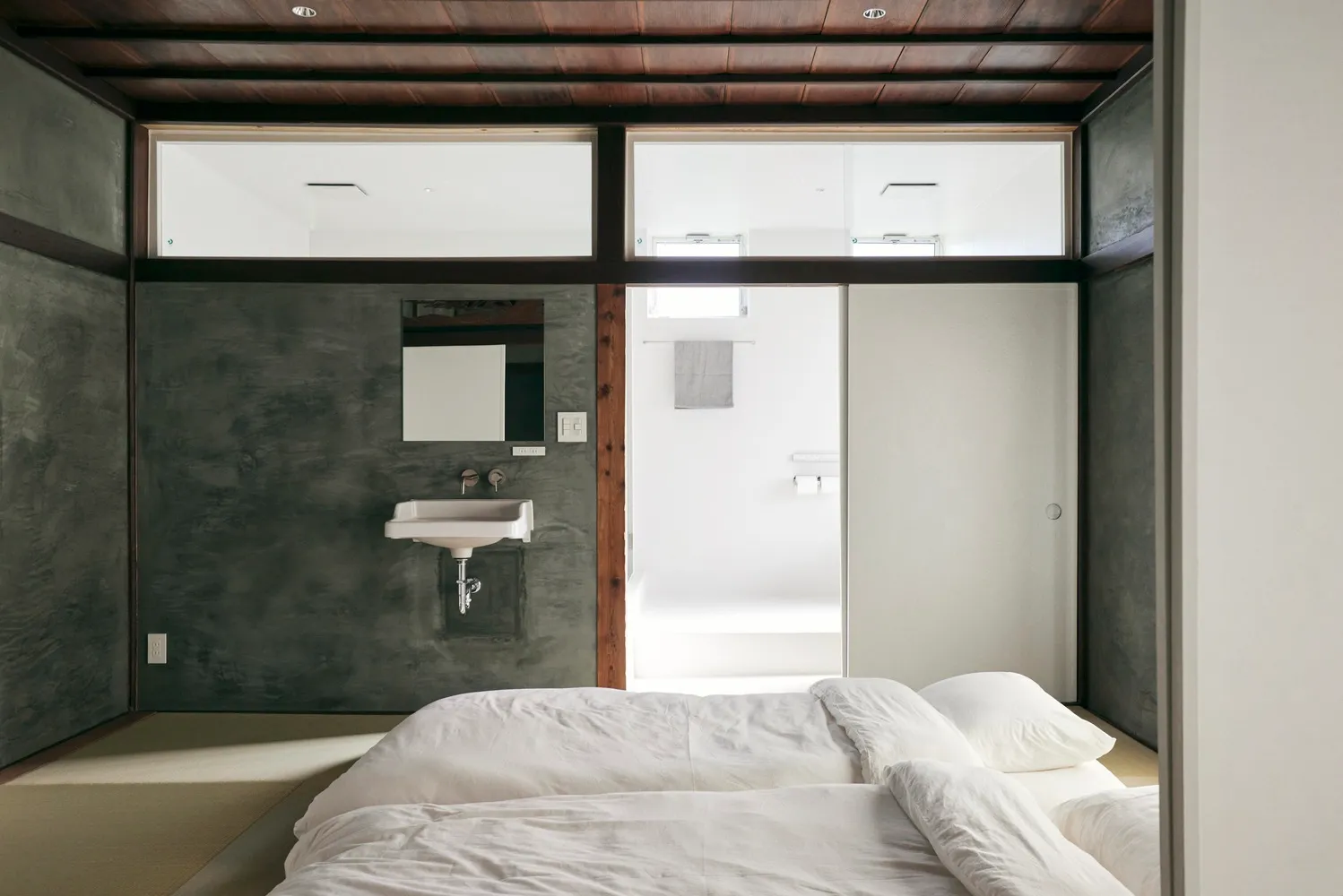 A minimalist bedroom with a concrete wall and floor, featuring a white bed and sink.