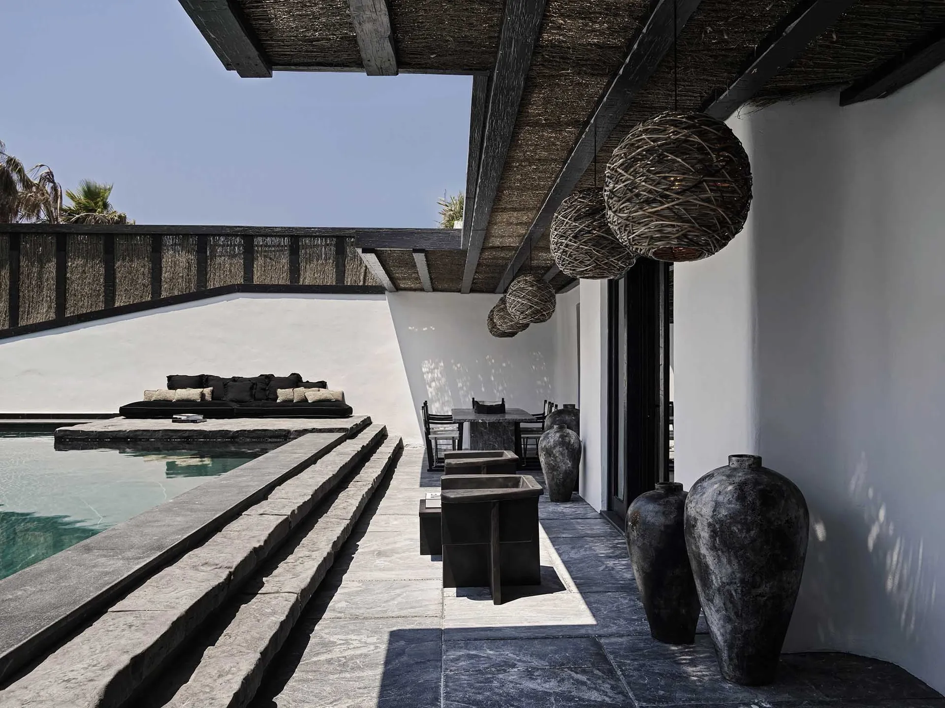 A modern minimalist outdoor living area featuring concrete and stone surfaces, rattan pendant lights, and a poolside seating
