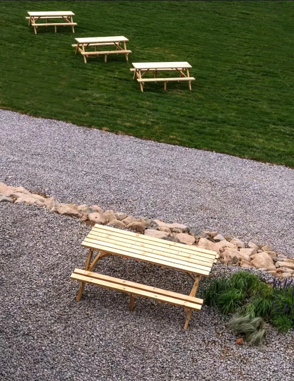 Four wooden picnic benches placed outdoors on a gravel surface surrounded by rocks and grass.
