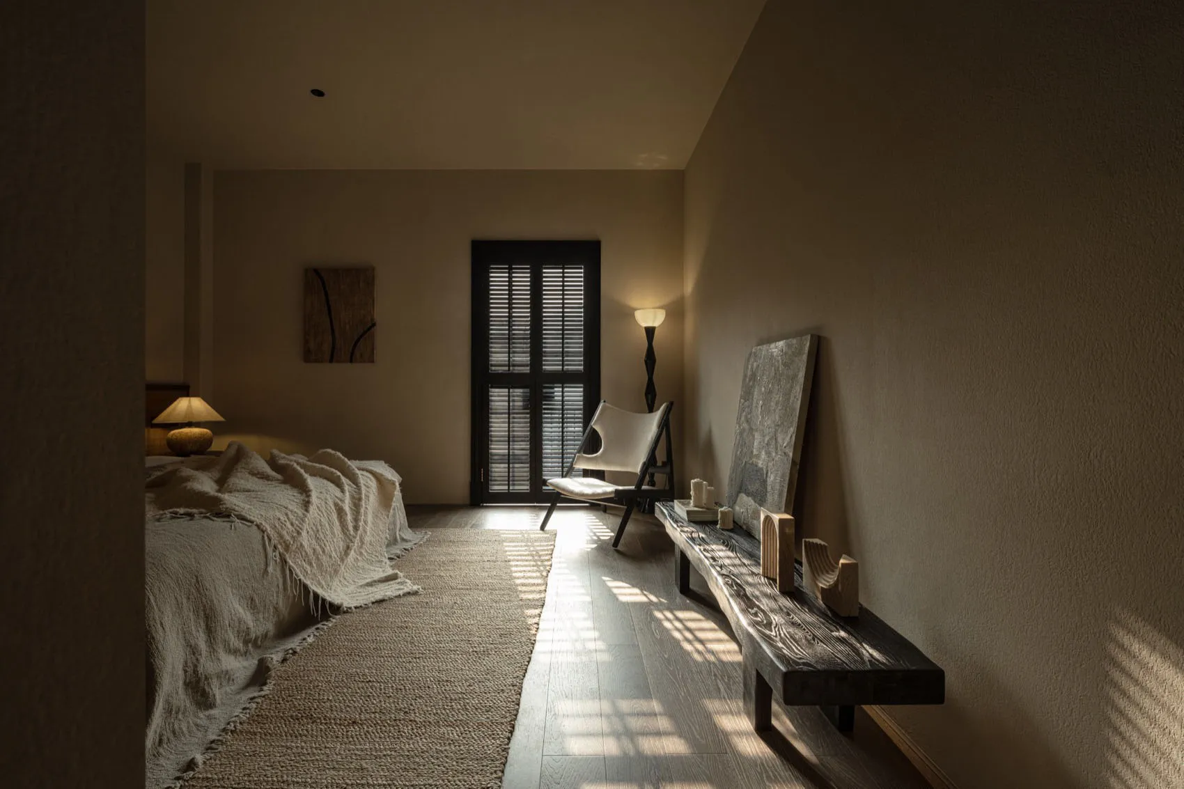 A minimalist bedroom with a bed covered in a textured blanket, a modern chair, and a wooden bench against a light-colored wal