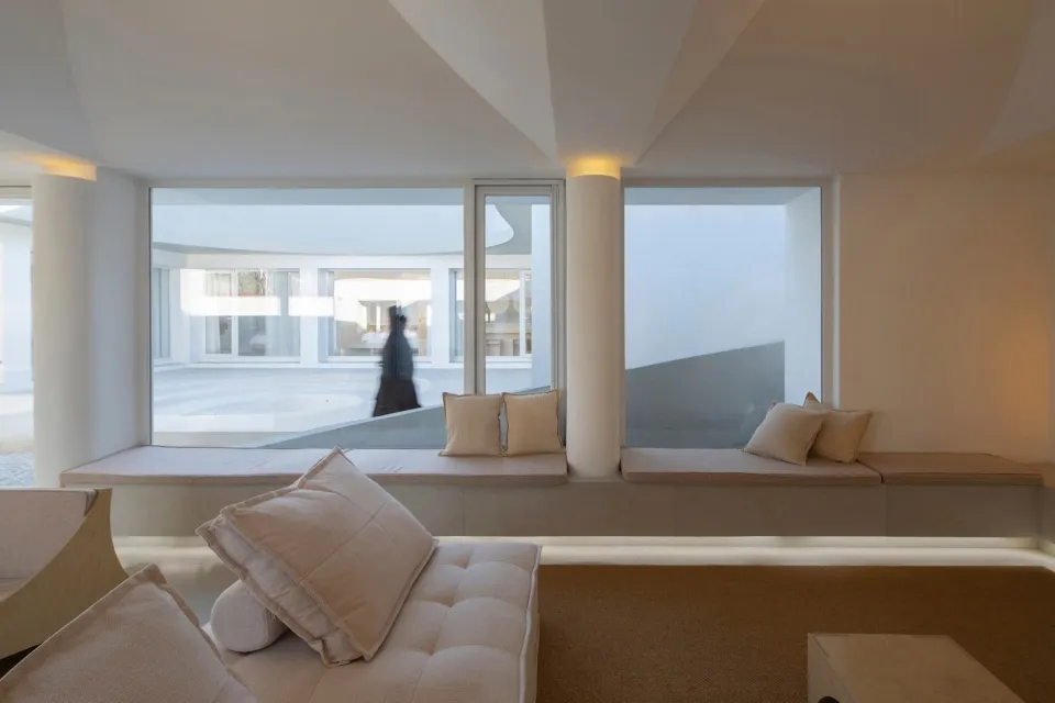 A minimalist living room with neutral-toned couches and cushions, large windows offering a view of an exterior space.