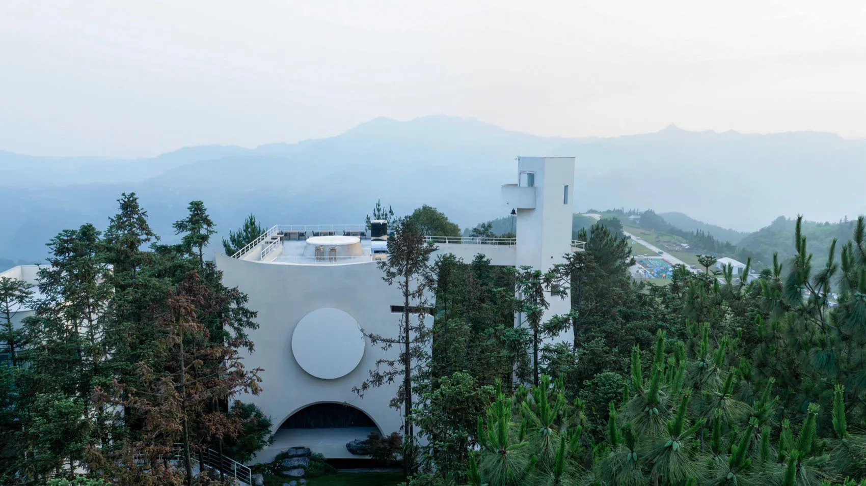 一棟現代白色住宅建築,設有白色圓窗與玻璃玄關門,周圍環繞綠意。