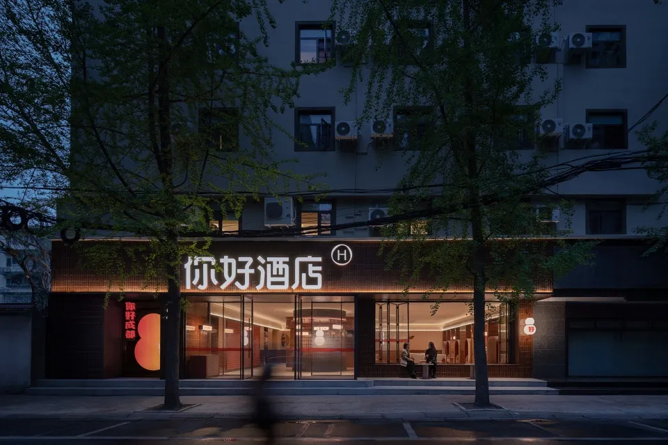 The storefront of a hotel featuring glass walls, concrete elements, and illuminated lighting fixtures at dusk.