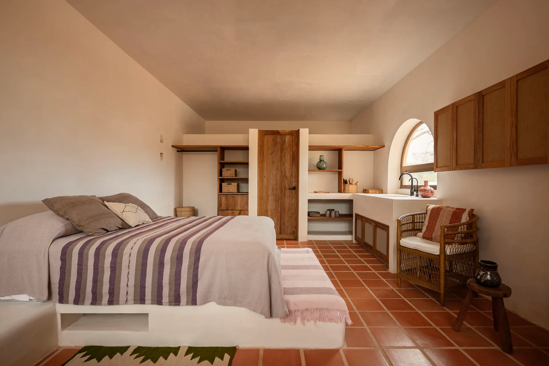A minimalist bedroom with terracotta tiles features a bed dressed in striped textiles, a wicker chair with striped cushions,