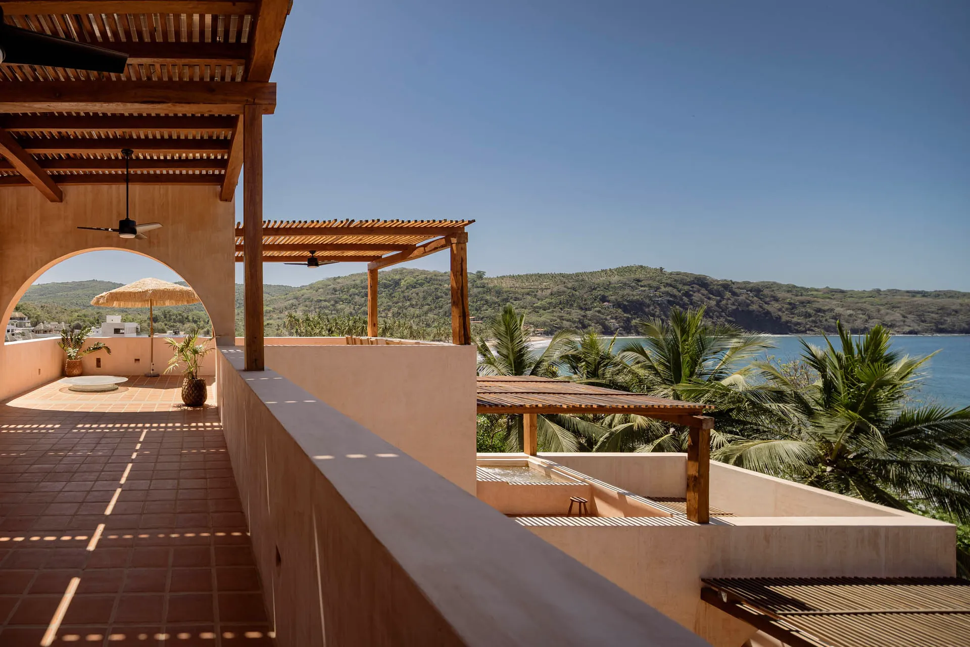 A spacious balcony featuring wood pergolas and concrete walls overlooking a lush tropical landscape and ocean.
