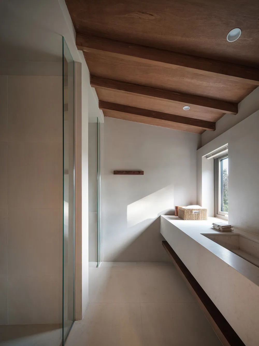 A minimalist bathroom with concrete walls and floor, featuring a wooden ceiling beam and a large window letting in natural li