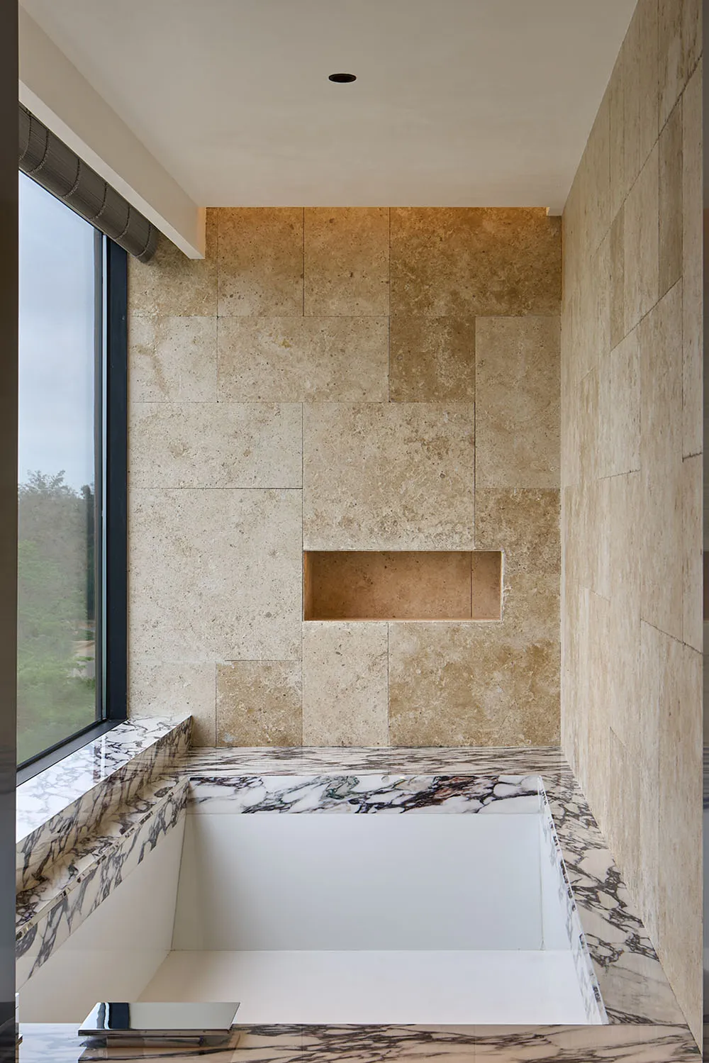 Modern, minimalist bathroom with a bathtub, marble floor, and travertine walls, featuring large window for natural light.