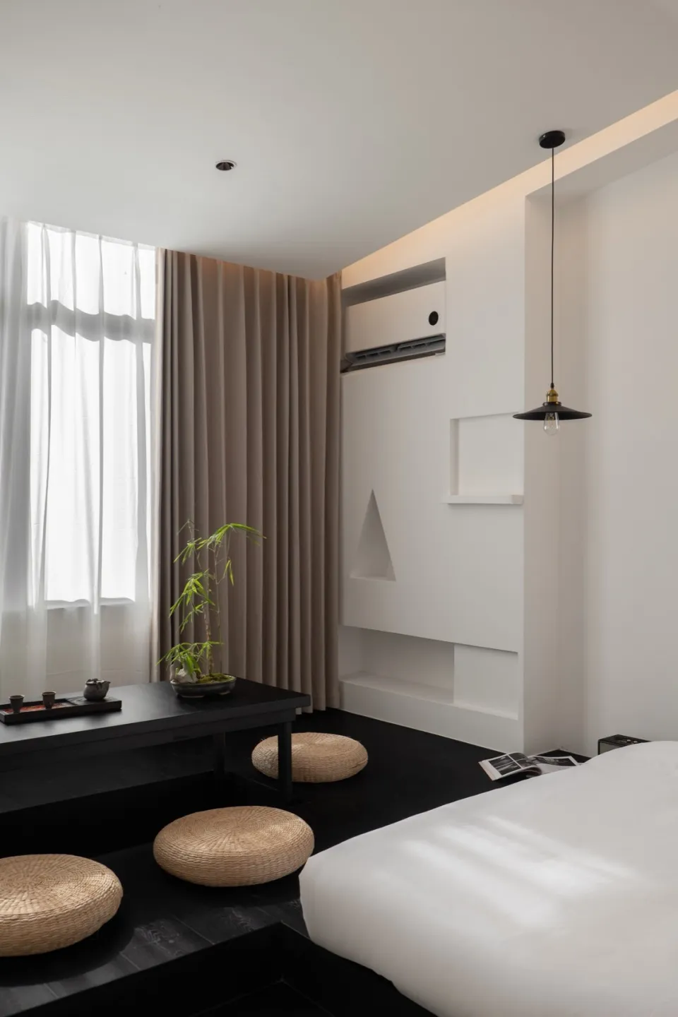 A modern minimalist bedroom with a white bed, wooden flooring, concrete walls, and floor cushions.