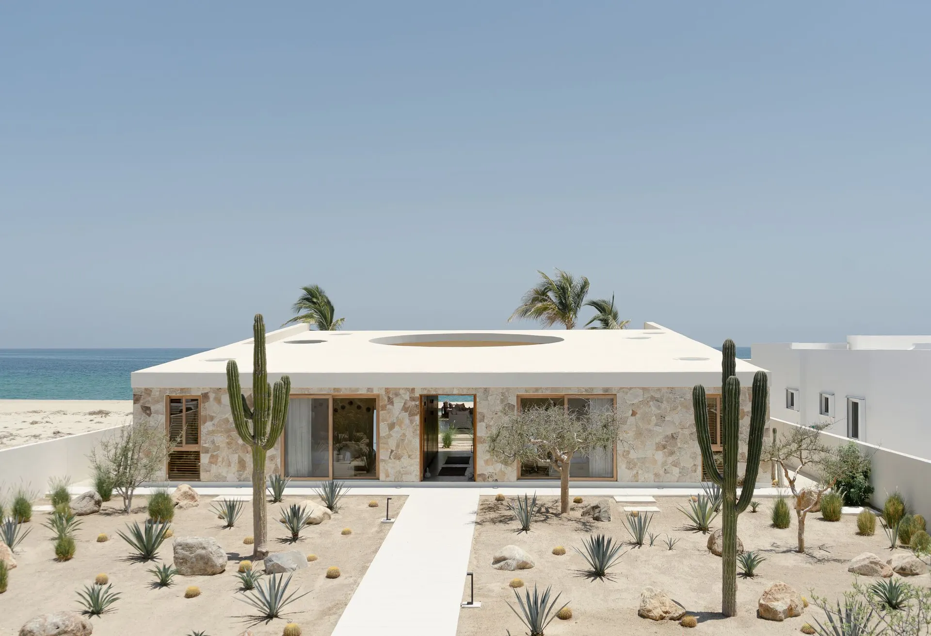 Modern building exterior with concrete walls and a circular cutout on the roof, set against a beach and ocean backdrop.
