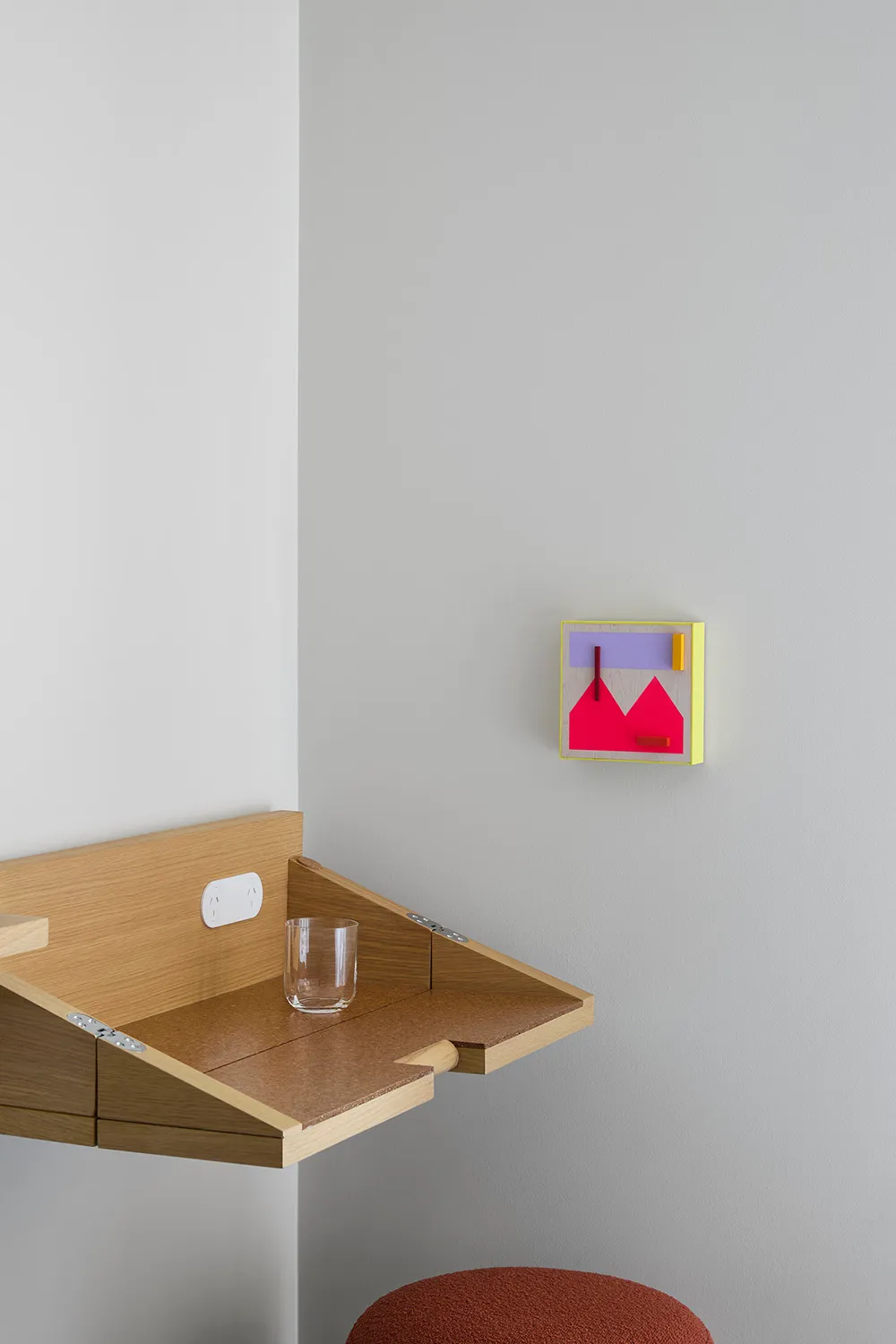 A minimalist home office space featuring a wooden desk with a glass surface, a glass tumbler, and a colorful abstract wall ar