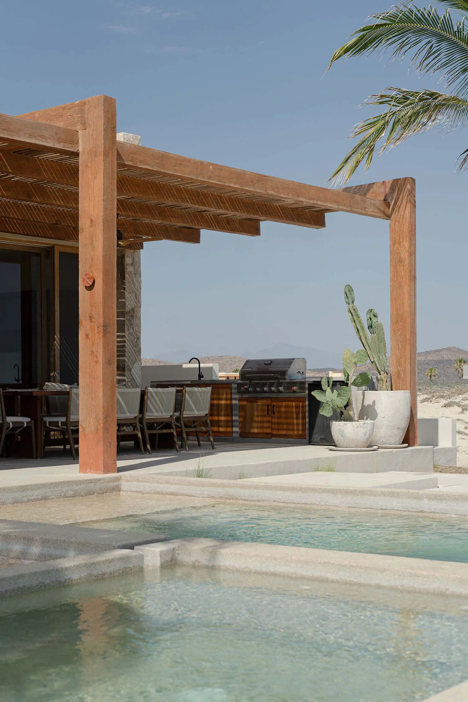 Modern residential pool area featuring a pergola-covered outdoor kitchen, grill, dining table, and cactus plants.