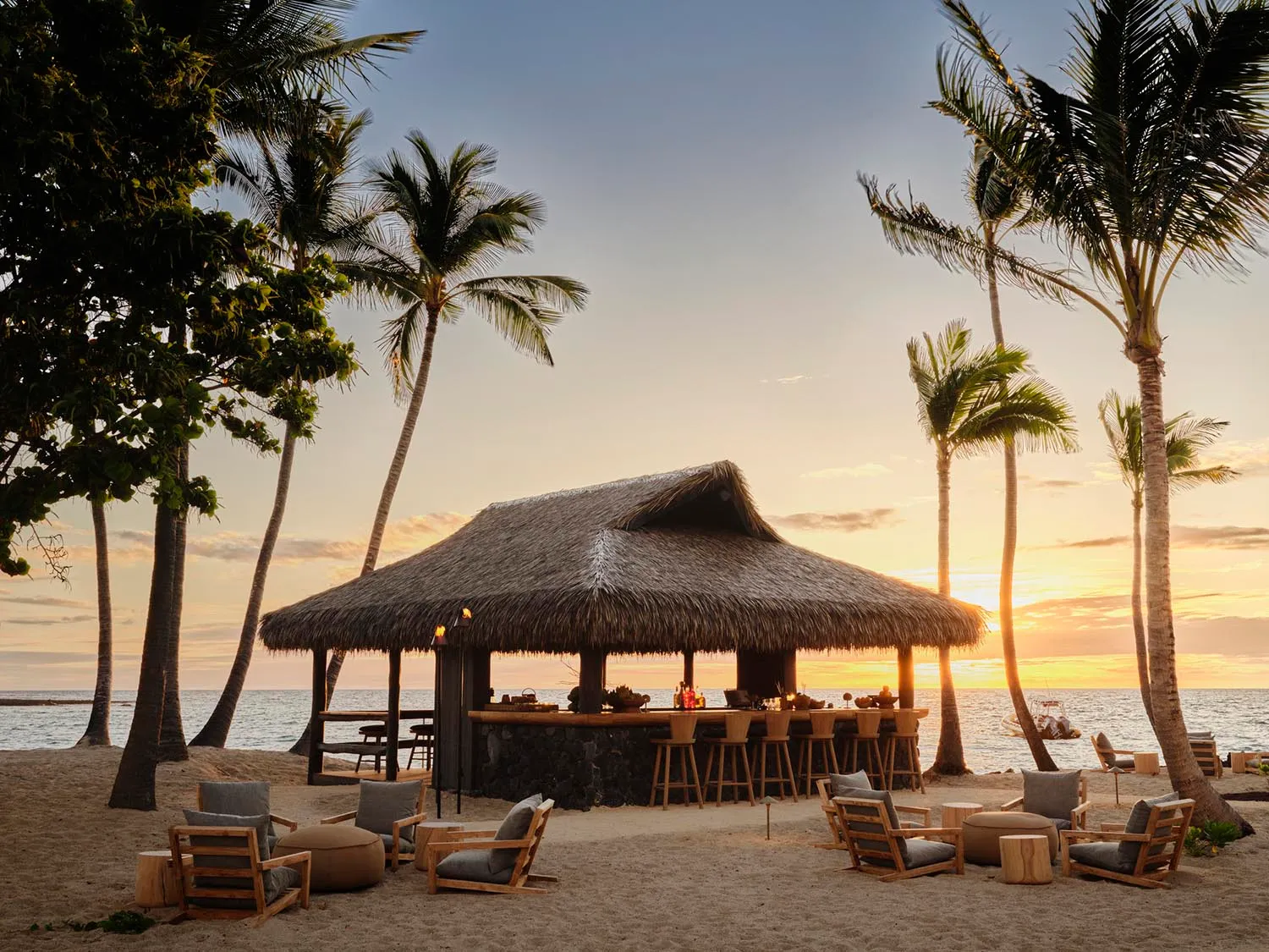 Thatched-roof beachside tiki bar with stools and lounge chairs under palm trees at sunset