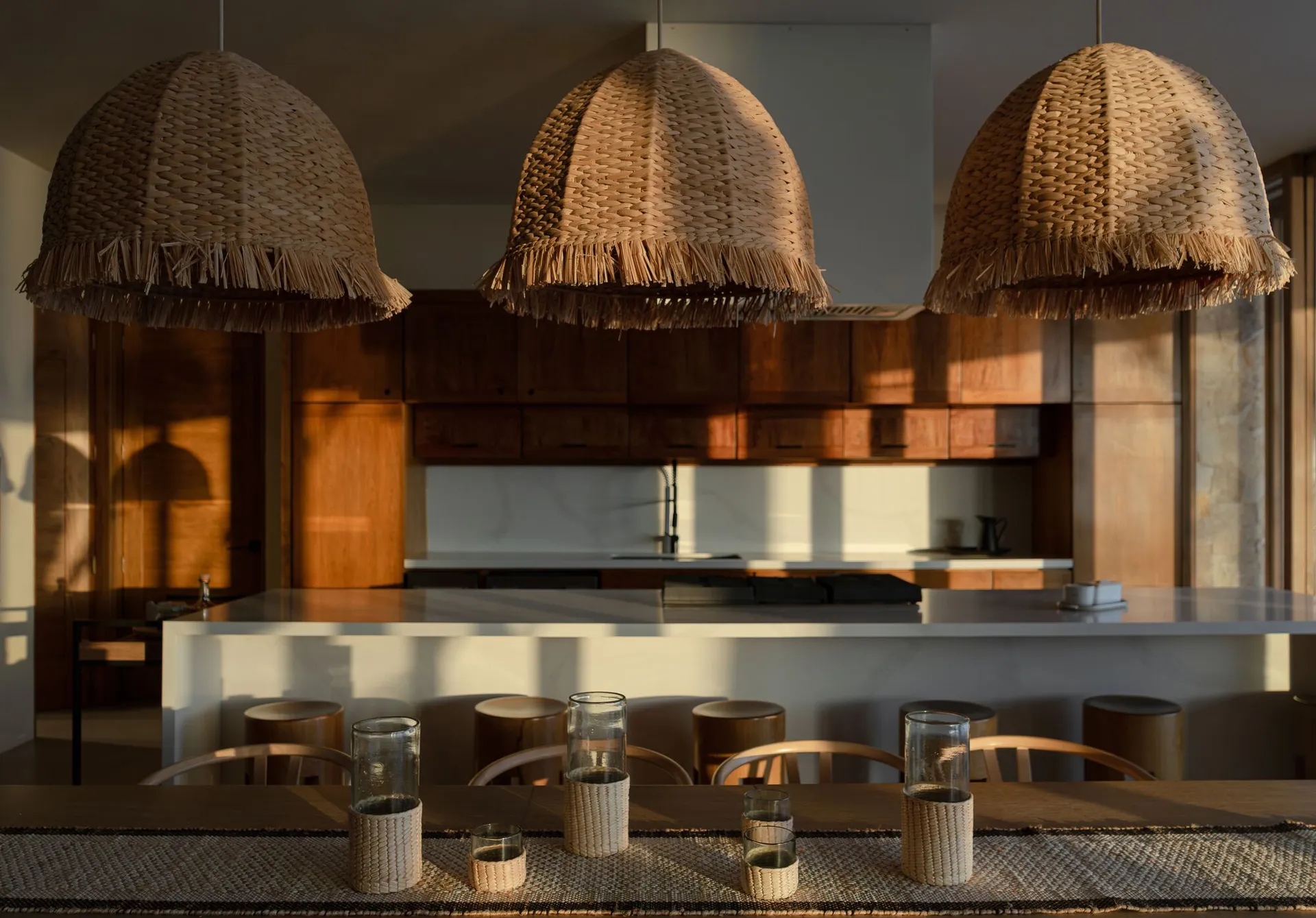 A modern kitchen featuring natural fiber lighting fixtures and wooden stools.