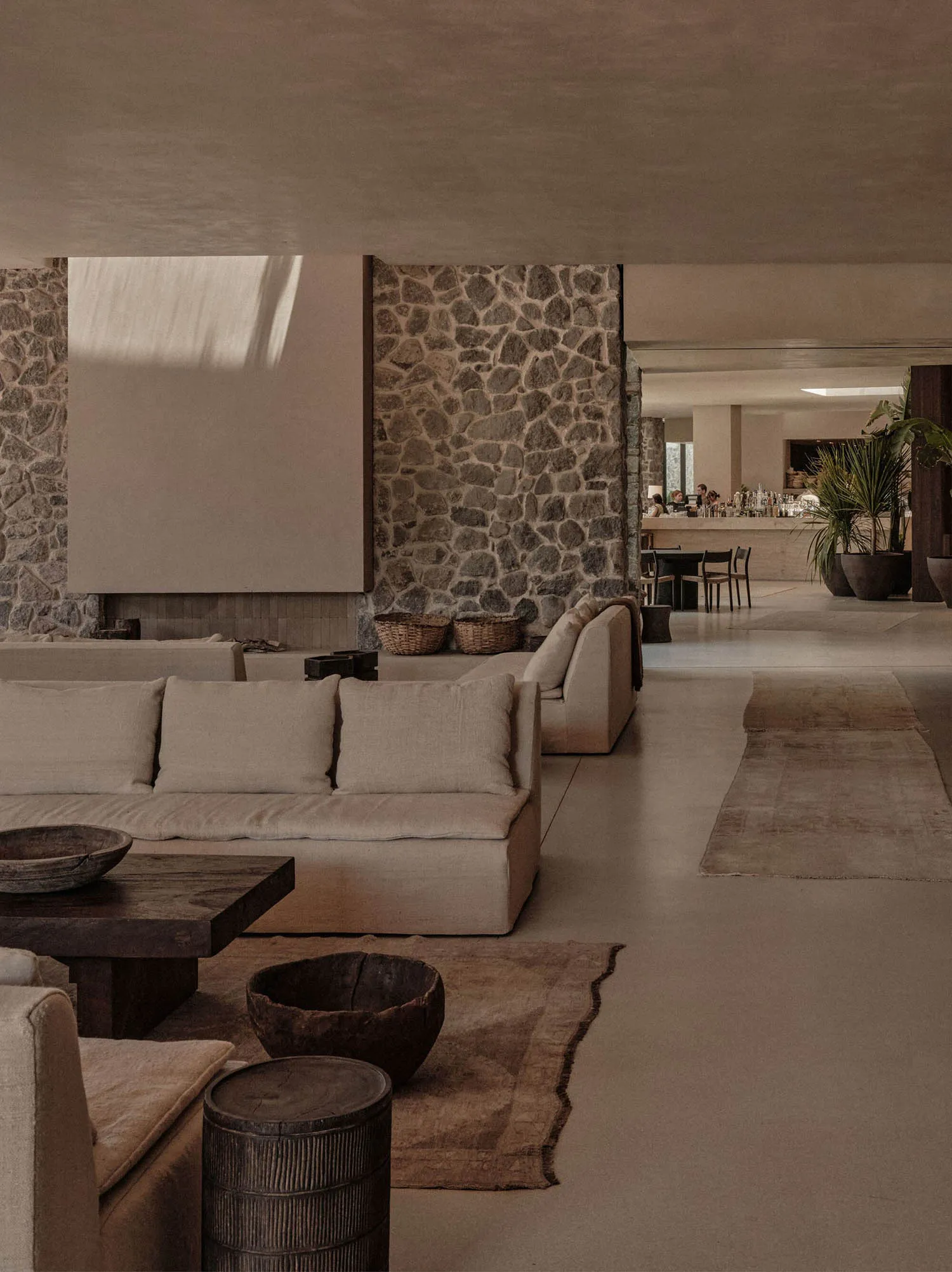 A minimalist lounge area with concrete furniture and flooring, featuring a stone wall and natural light.