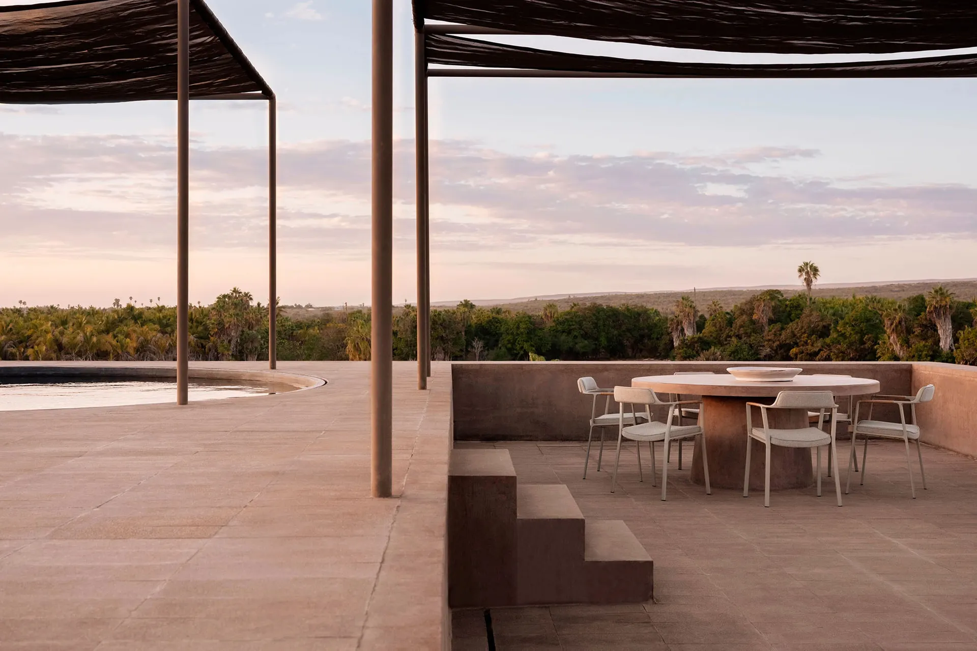 A modern outdoor lounge space with concrete flooring and furniture, surrounded by nature.