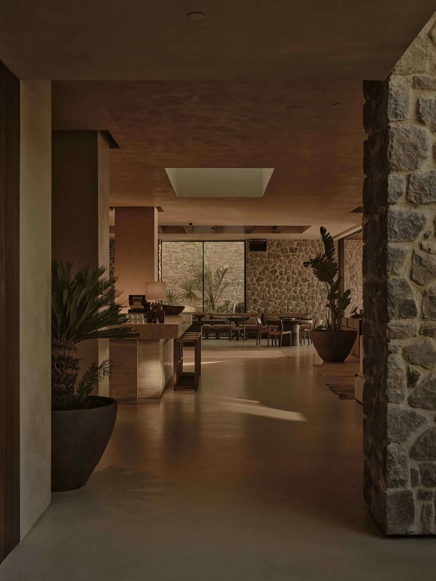 A modern living room with concrete flooring and walls, featuring a large stone fireplace and potted plants.