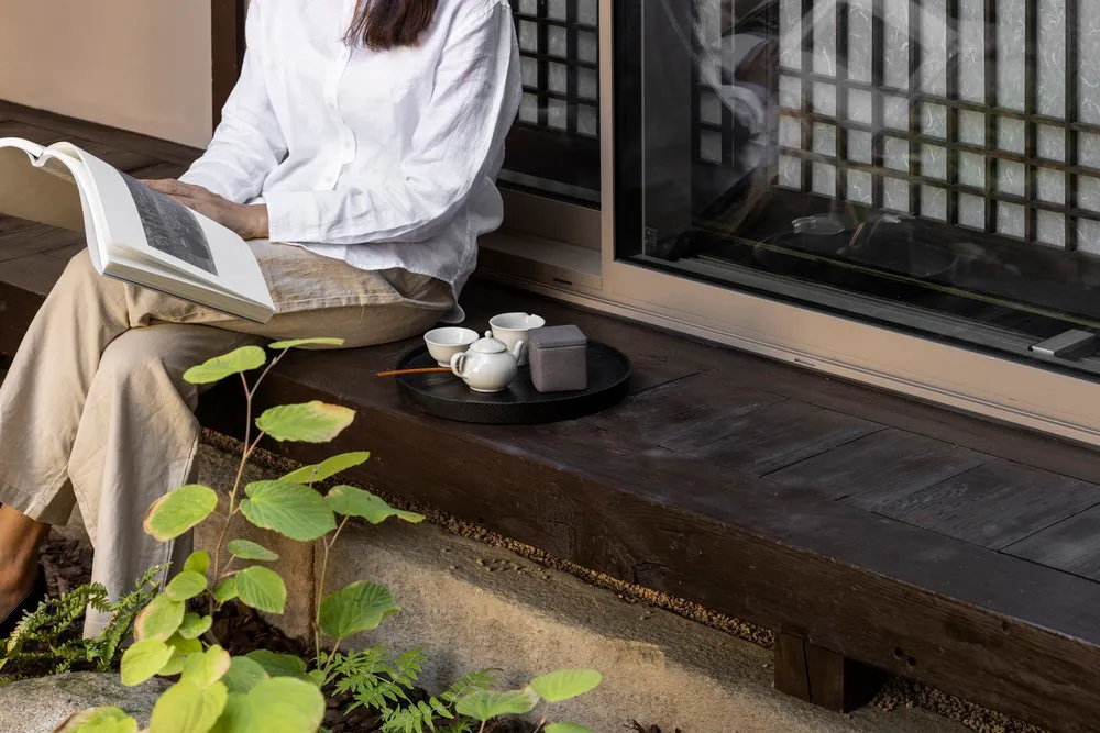 Person sitting on a wooden floor with a tea set and reading a book.