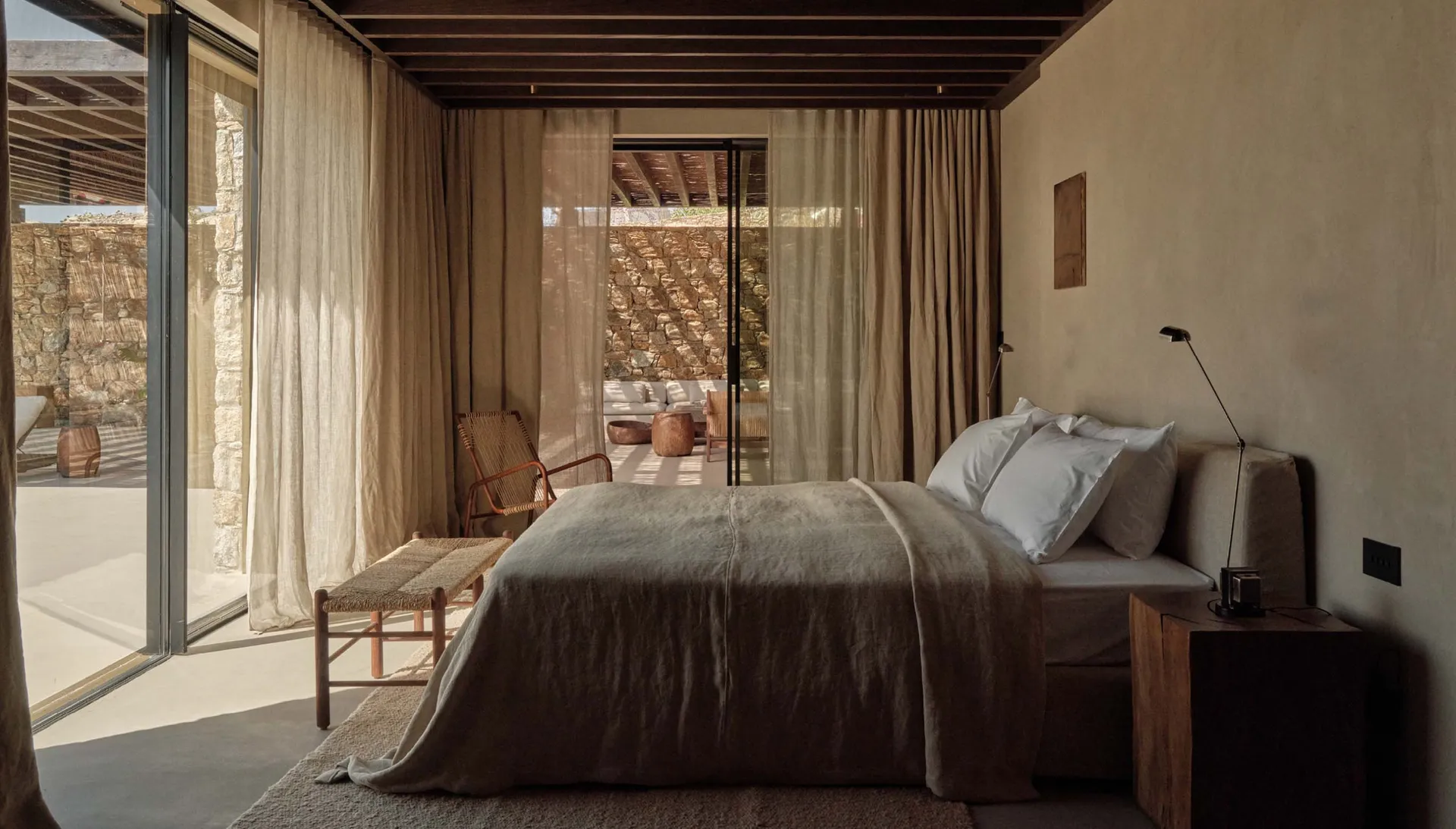 A minimalist bedroom with linen bedding, wooden furniture, and a stone wall, featuring a view blocked by curtains.
