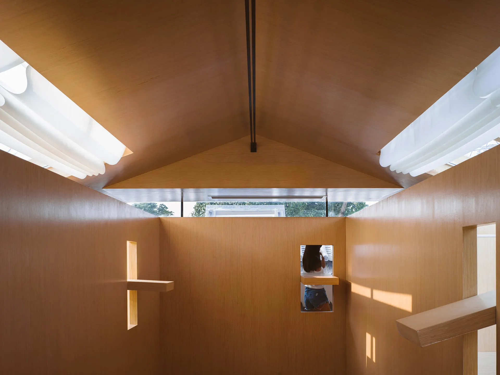 A minimalist interior with wooden walls and a sloped wooden ceiling, featuring a small window and minimalist lighting fixture