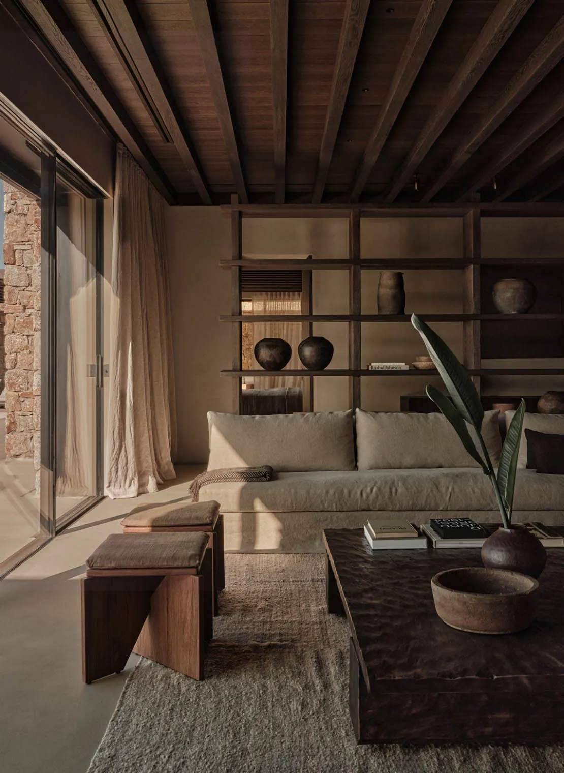 A modern, minimalist living room with wooden furniture, stone walls, and a large window allowing natural light to illuminate