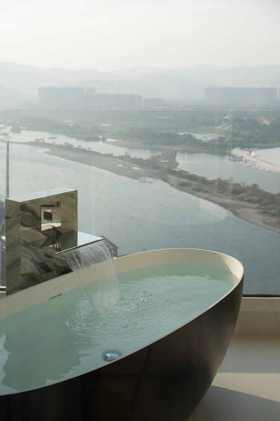 Modern bathroom with glass waterfall bathtub overlooking scenic river and cityscape.
