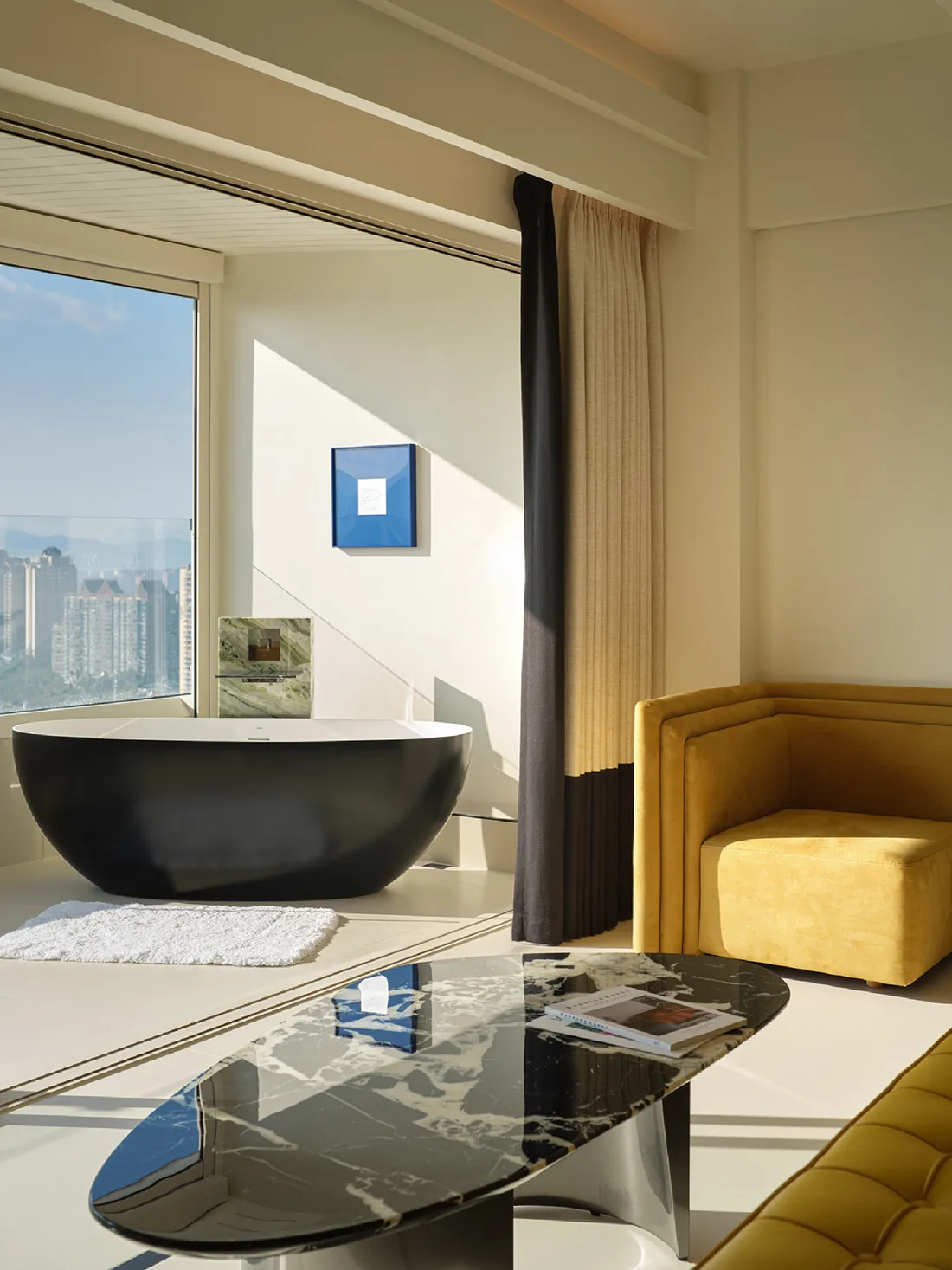 A modern suite bathroom featuring a black freestanding bathtub and a living area with a yellow armchair and coffee table.
