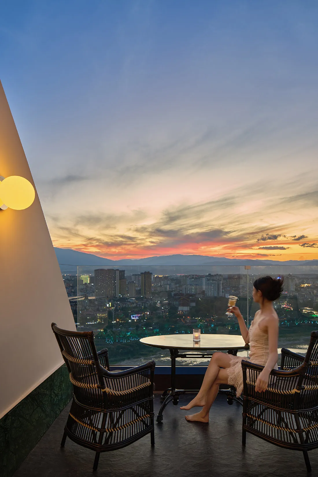 A woman sits in a wicker chair on a rooftop, enjoying a drink at sunset with a cityscape view.