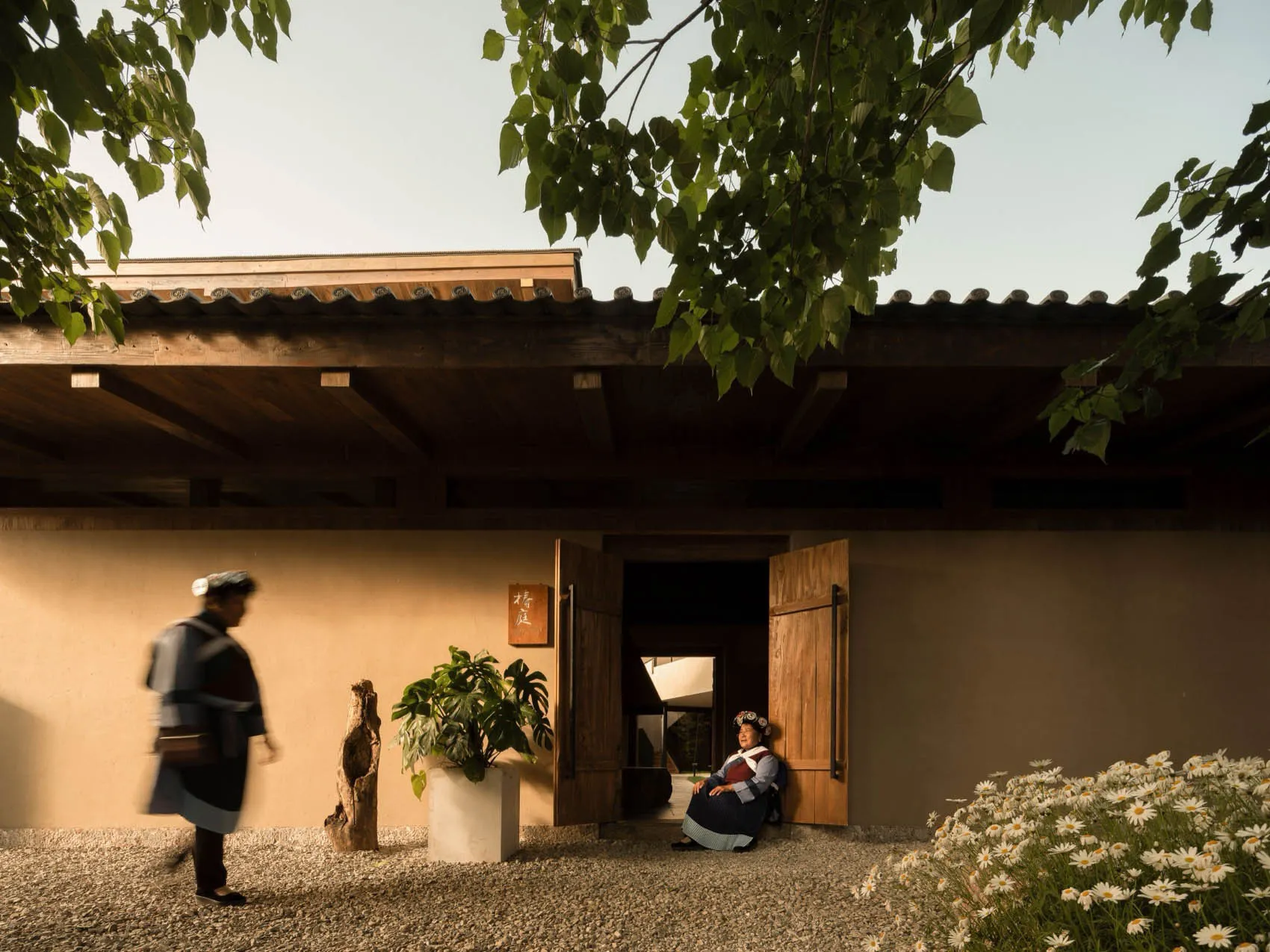 A traditional wooden residence with a wooden roof and walls, featuring a person sitting at the entrance and white flowers in