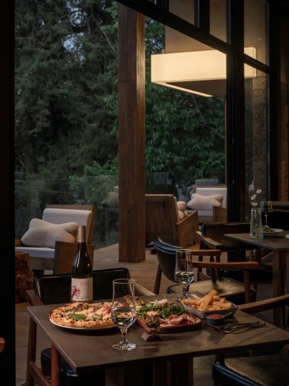 A wooden restaurant interior featuring a table with food and wine, chairs, and a window with a view of trees.