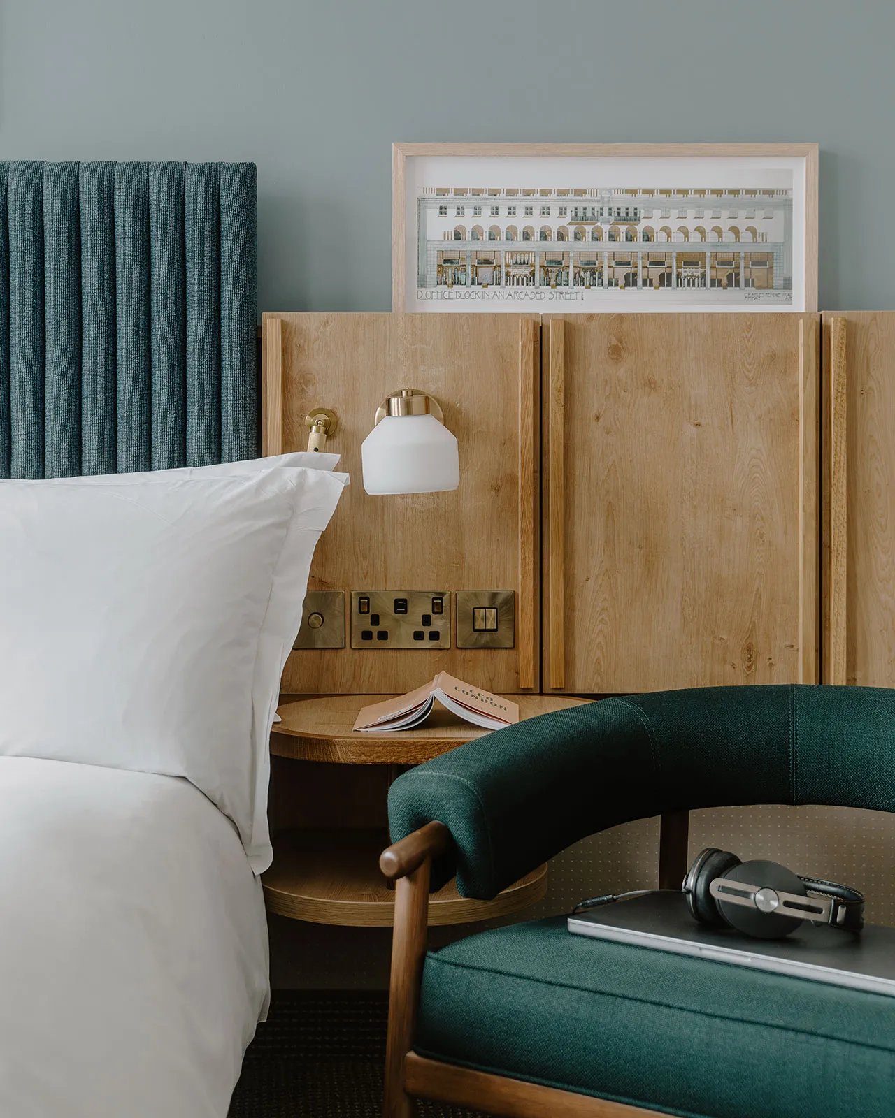 A minimalist hotel room featuring a bed with a wooden headboard and a wooden nightstand with a lamp, a chair with a wooden ar