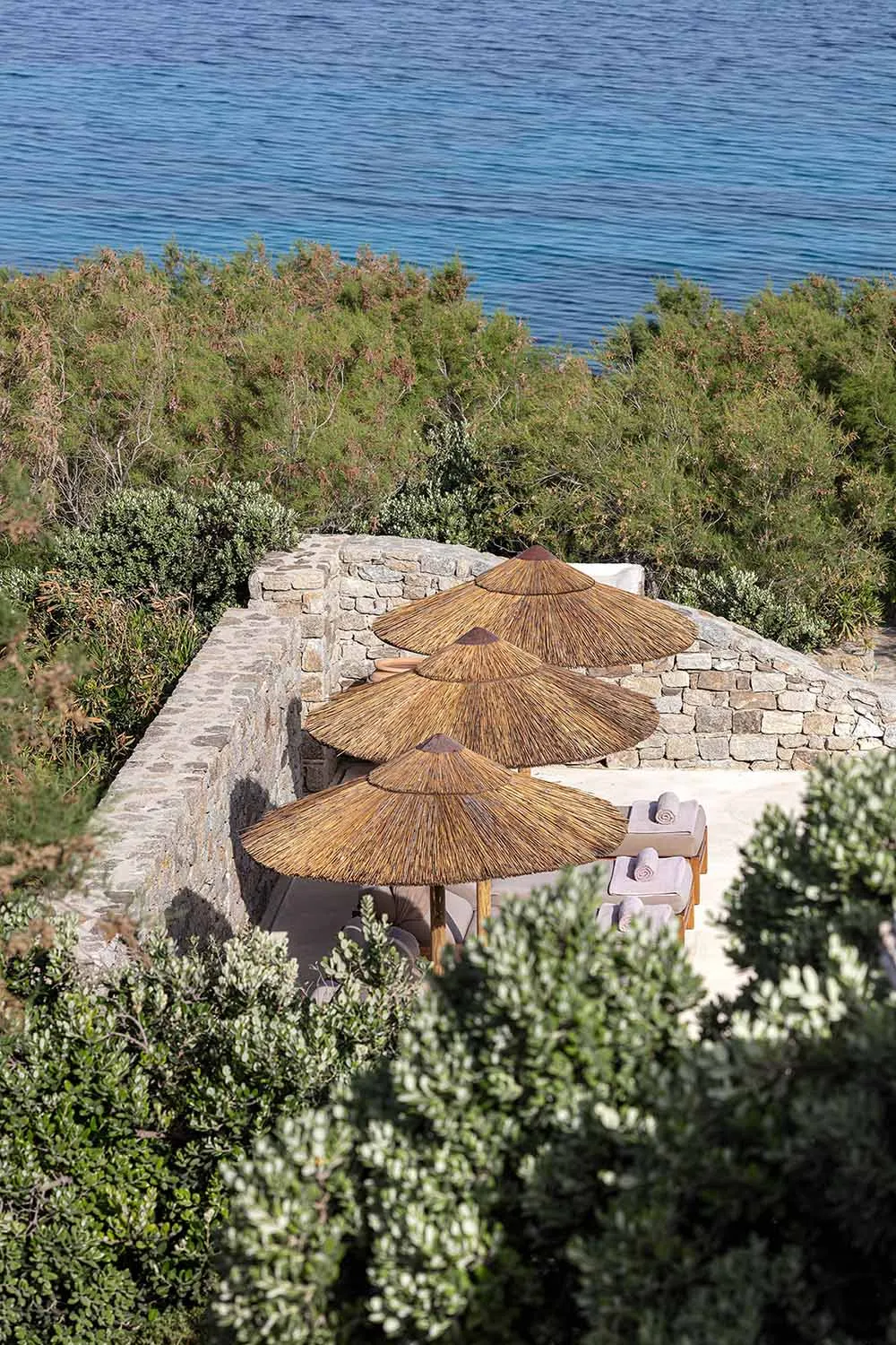 Three thatched umbrellas and sun loungers on a stone terrace overlooking the ocean and greenery.