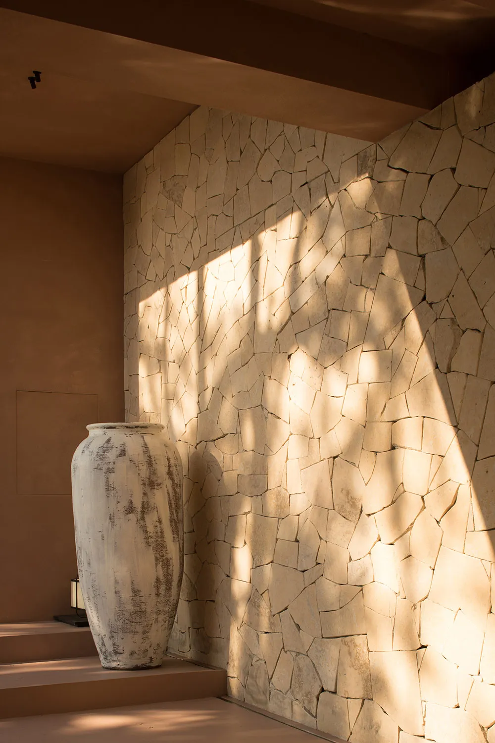 A close-up view of a corner featuring a textured stone wall with a large, weathered vase on a step, casting shadows and illum