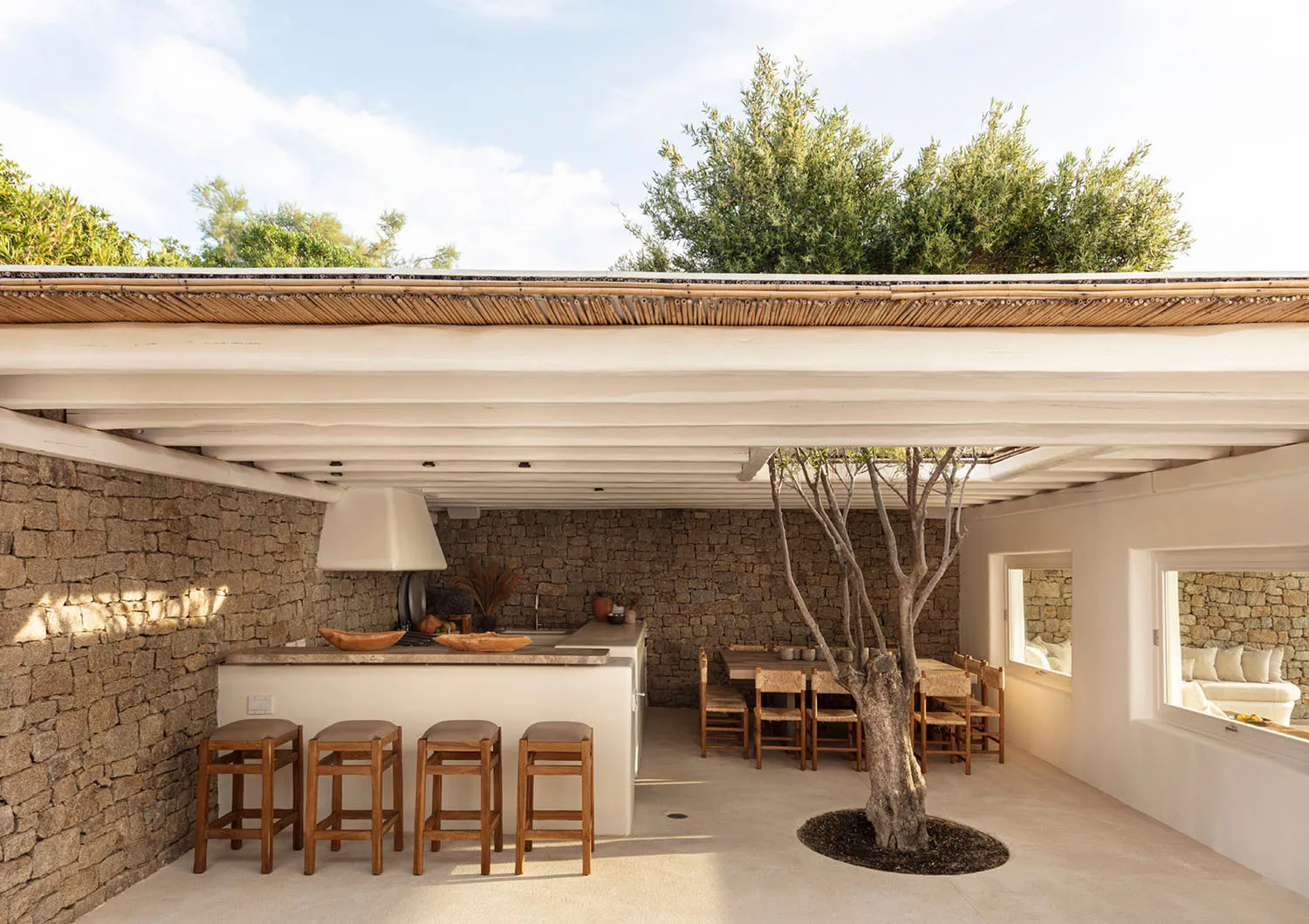 An outdoor kitchen area featuring a modern design with concrete countertops, stone walls, and timber stools and table.