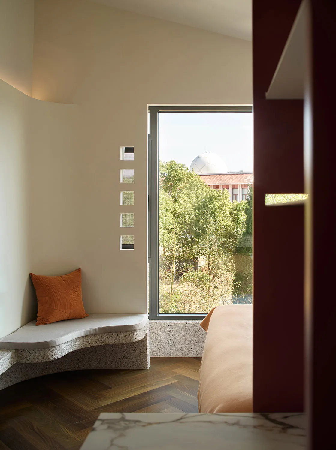 A minimalist bedroom featuring a concrete bench with a cushion and a window overlooking trees and a building.