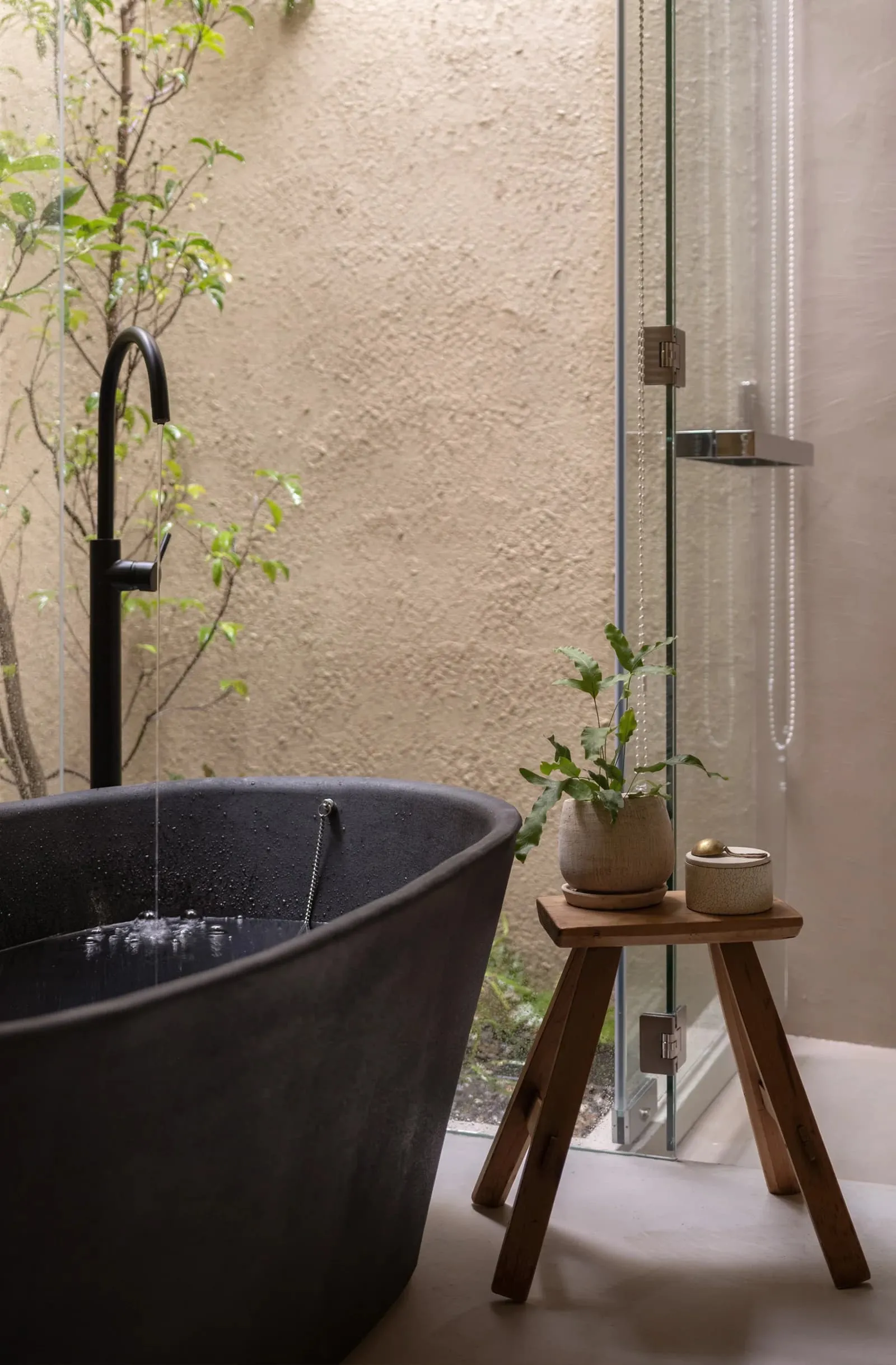 A modern, minimalist bathroom with a freestanding concrete bathtub, a small wooden stool with a plant and a container, and a