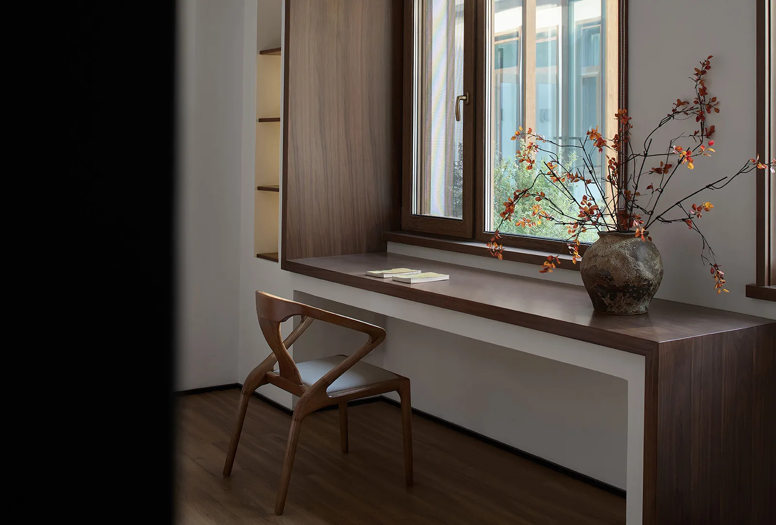A modern library space featuring a wooden desk, chair, and bookshelves with a vase of autumn leaves on the windowsill.