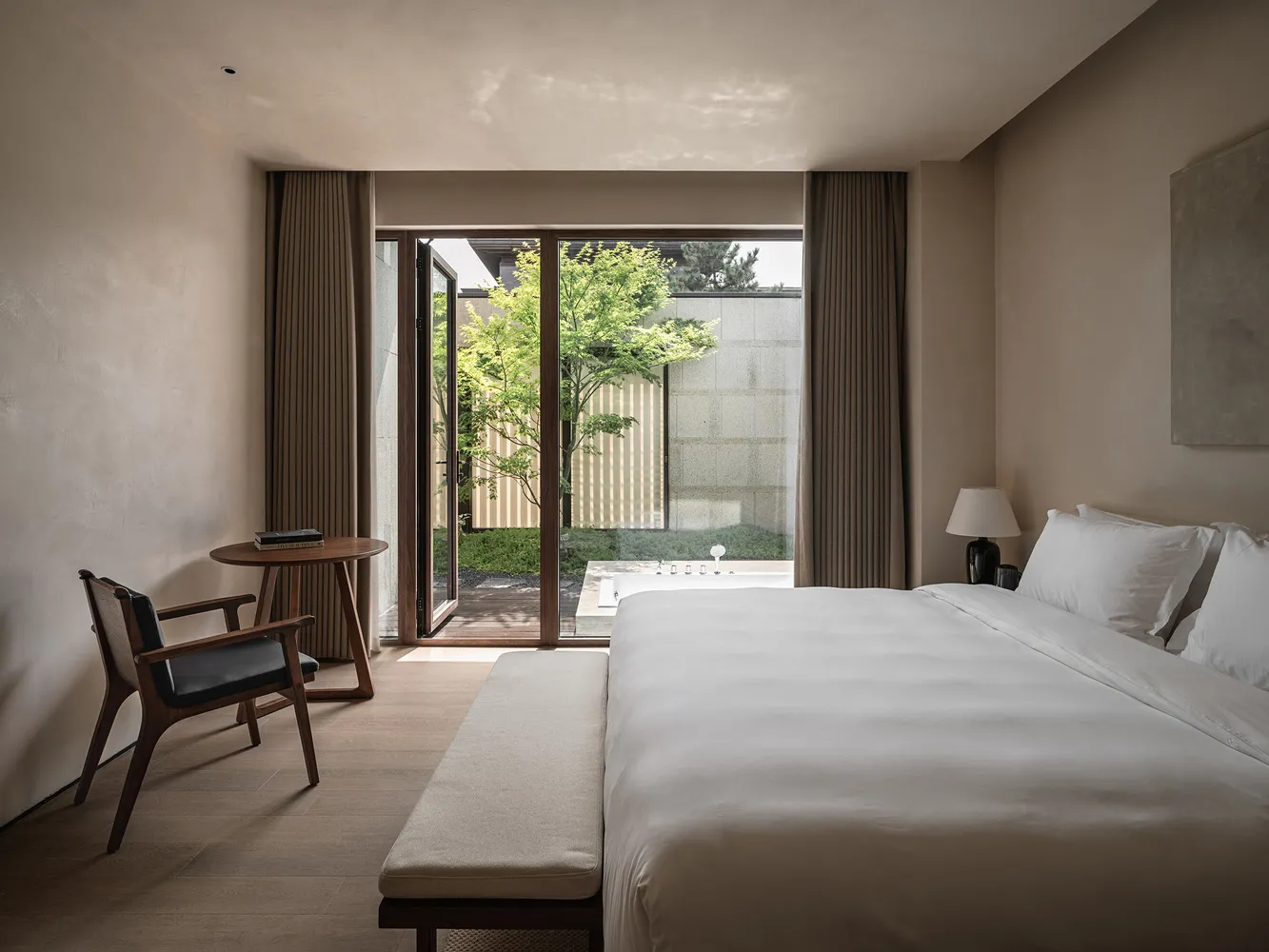 A minimalist bedroom with a white fabric bed on a wooden floor, featuring natural light and greenery outside through large wi
