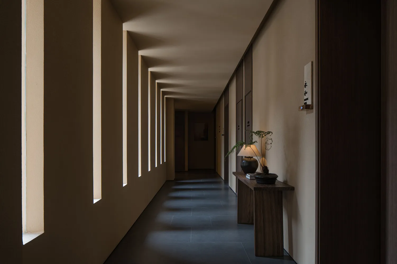 A minimalist corridor with concrete walls and floor, featuring rectangular windows and a wooden side table with a lamp and po