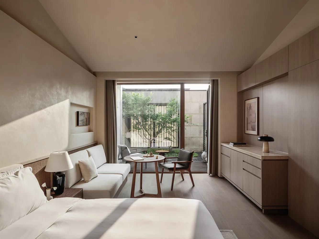 A minimalist bedroom featuring wooden furniture and textured textiles, with a view of greenery through large windows.