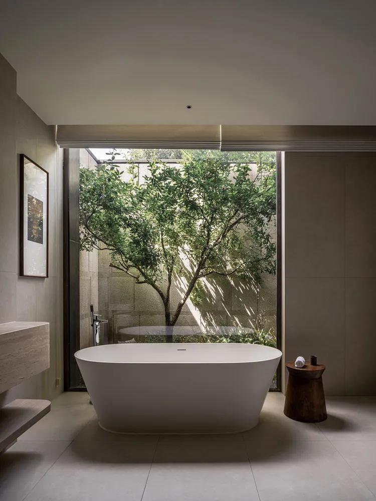 Modern minimalist bathroom featuring a freestanding bathtub with concrete walls and a large window overlooking a tree.