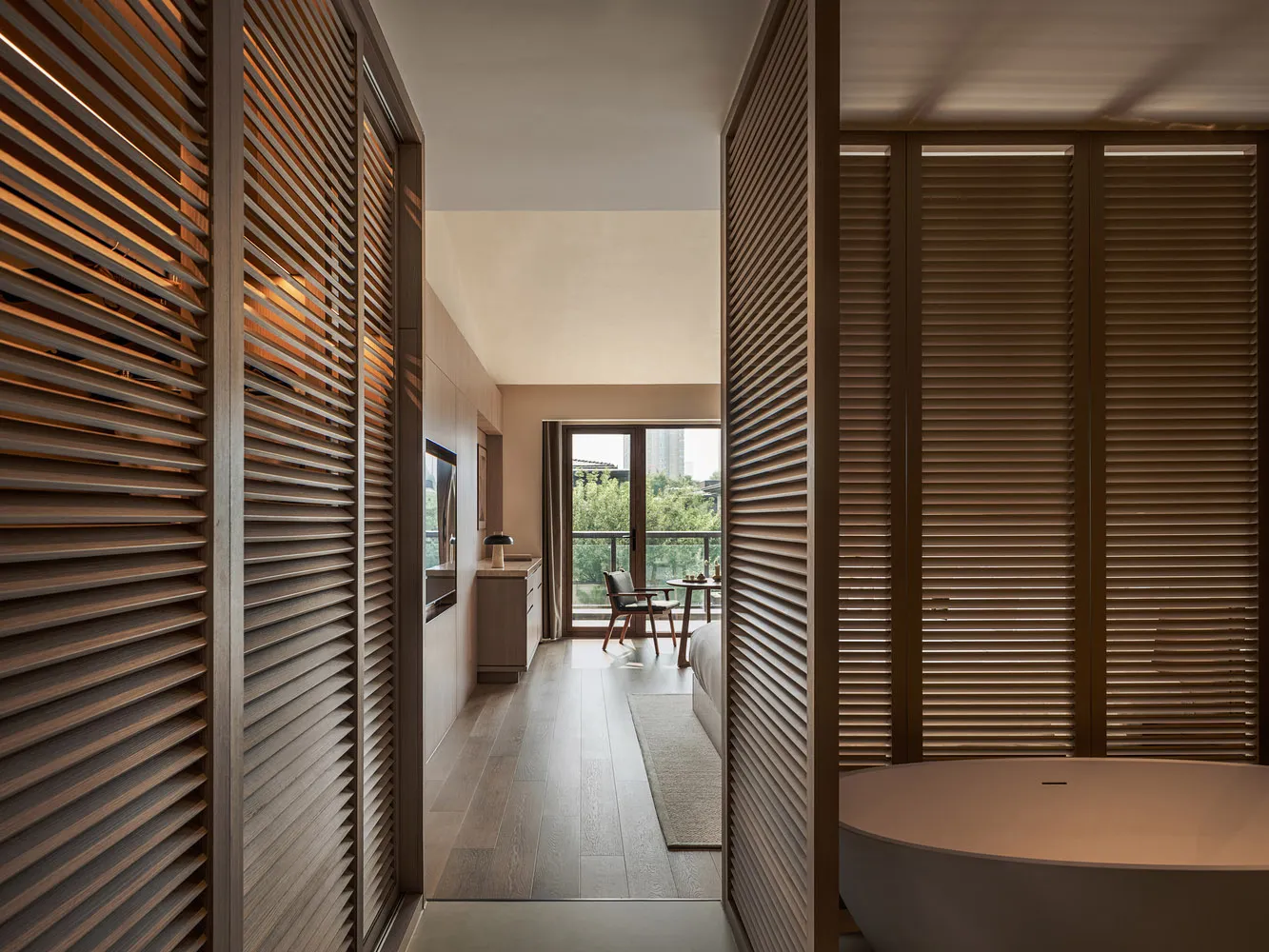 A minimalistic bedroom featuring wooden furniture and a freestanding bathtub, with a view of the outdoor balcony and greenery