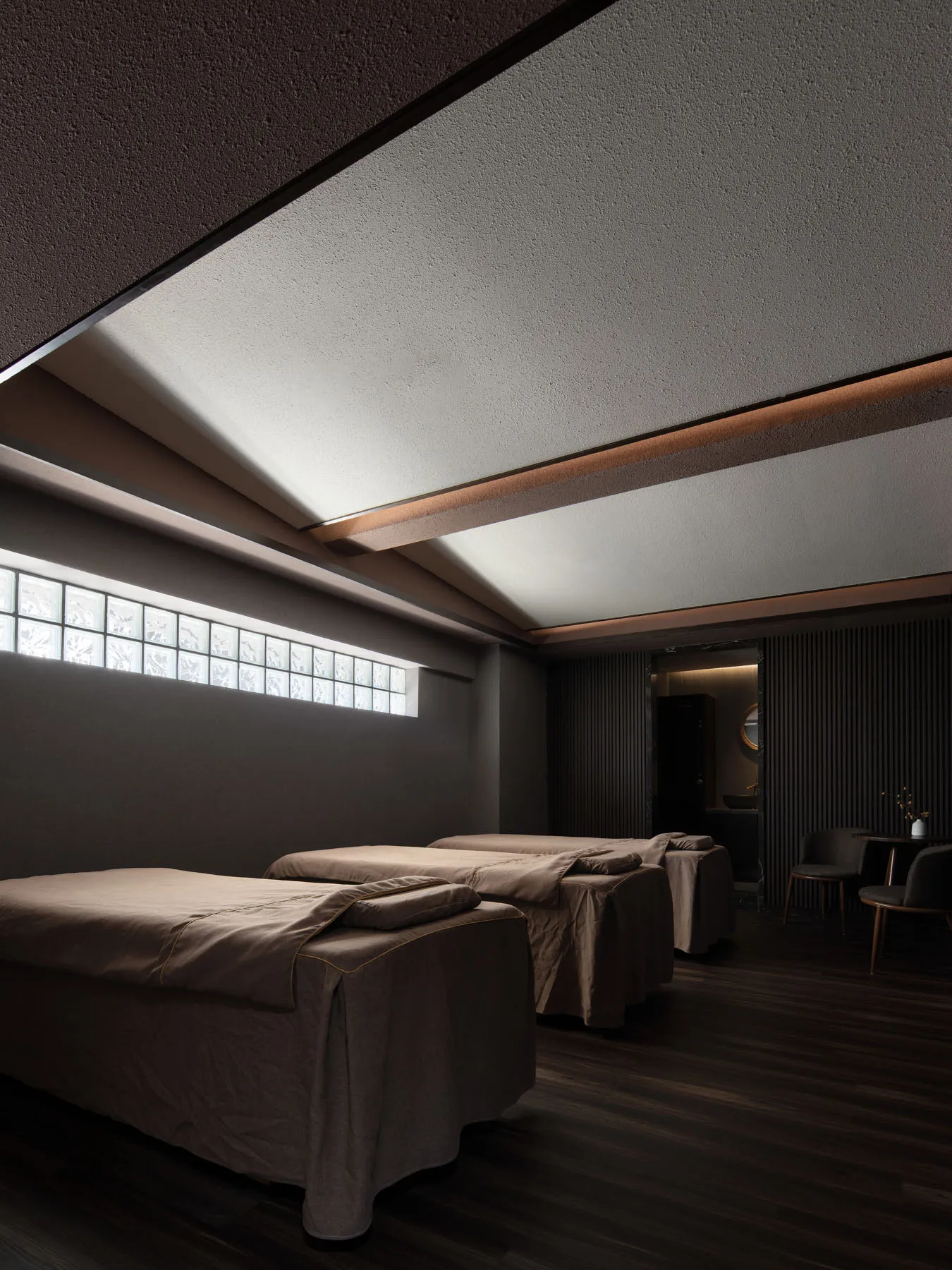 Spa room with three massage beds covered with brown fabric, dark interior, and small window with square glass panels.