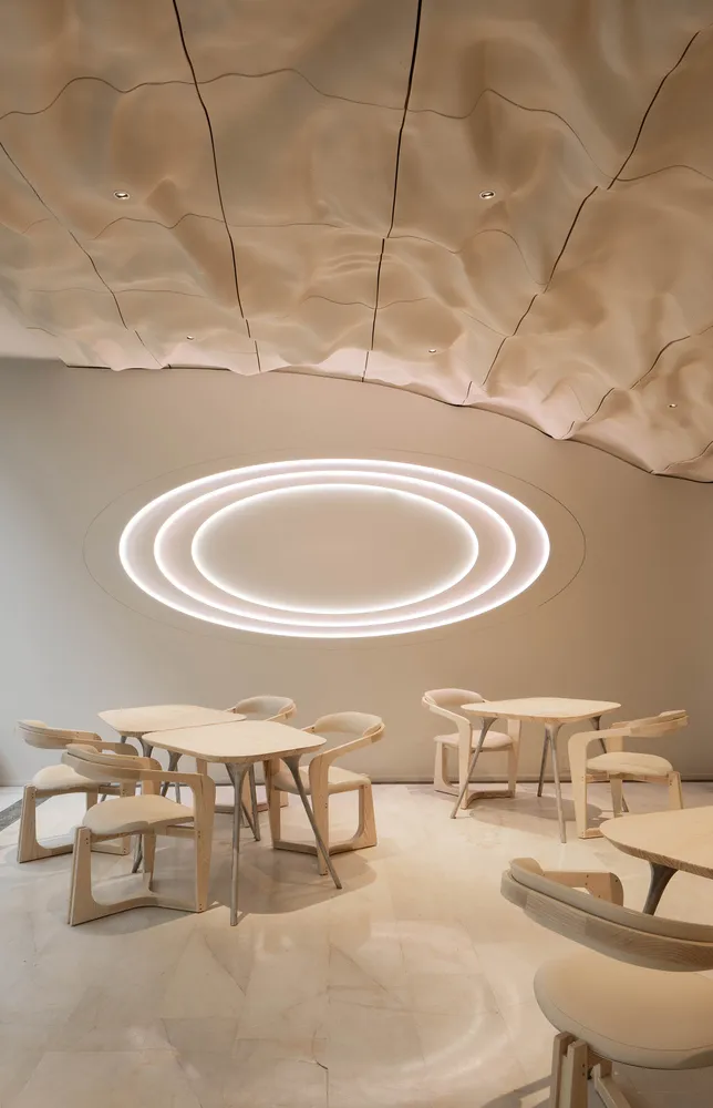 A minimalist restaurant with white walls, wooden tables, and chairs, illuminated by circular lights in the ceiling.
