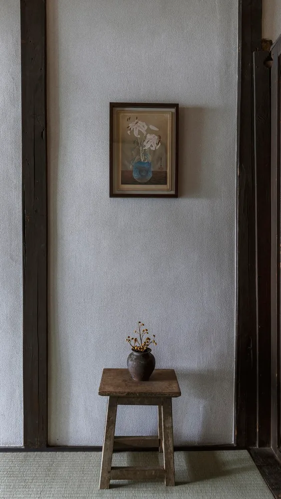 A minimalist studio featuring a small wooden stool with a vase of wilted flowers and a framed painting of flowers on the wall