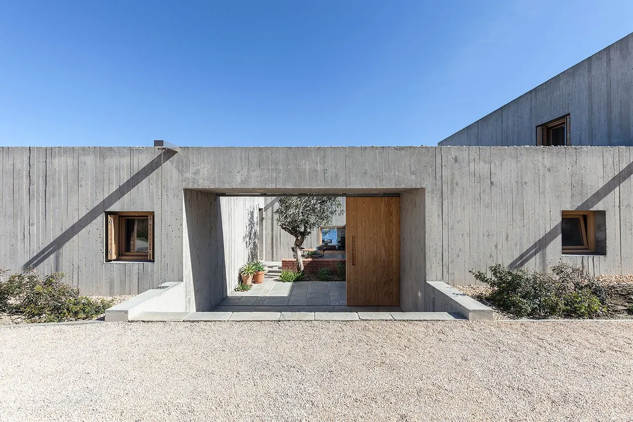 A modern concrete house with a wooden door and a small tree in front of it, set against a clear blue sky.