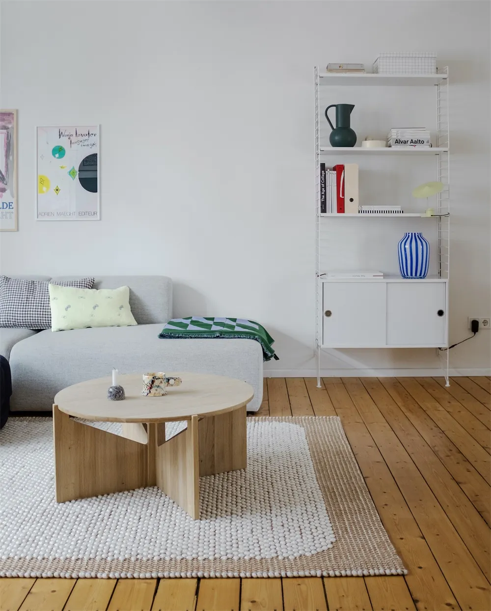 A modern minimalist living room featuring a light-colored oak wood coffee table, a grey couch, and a white wire shelving unit