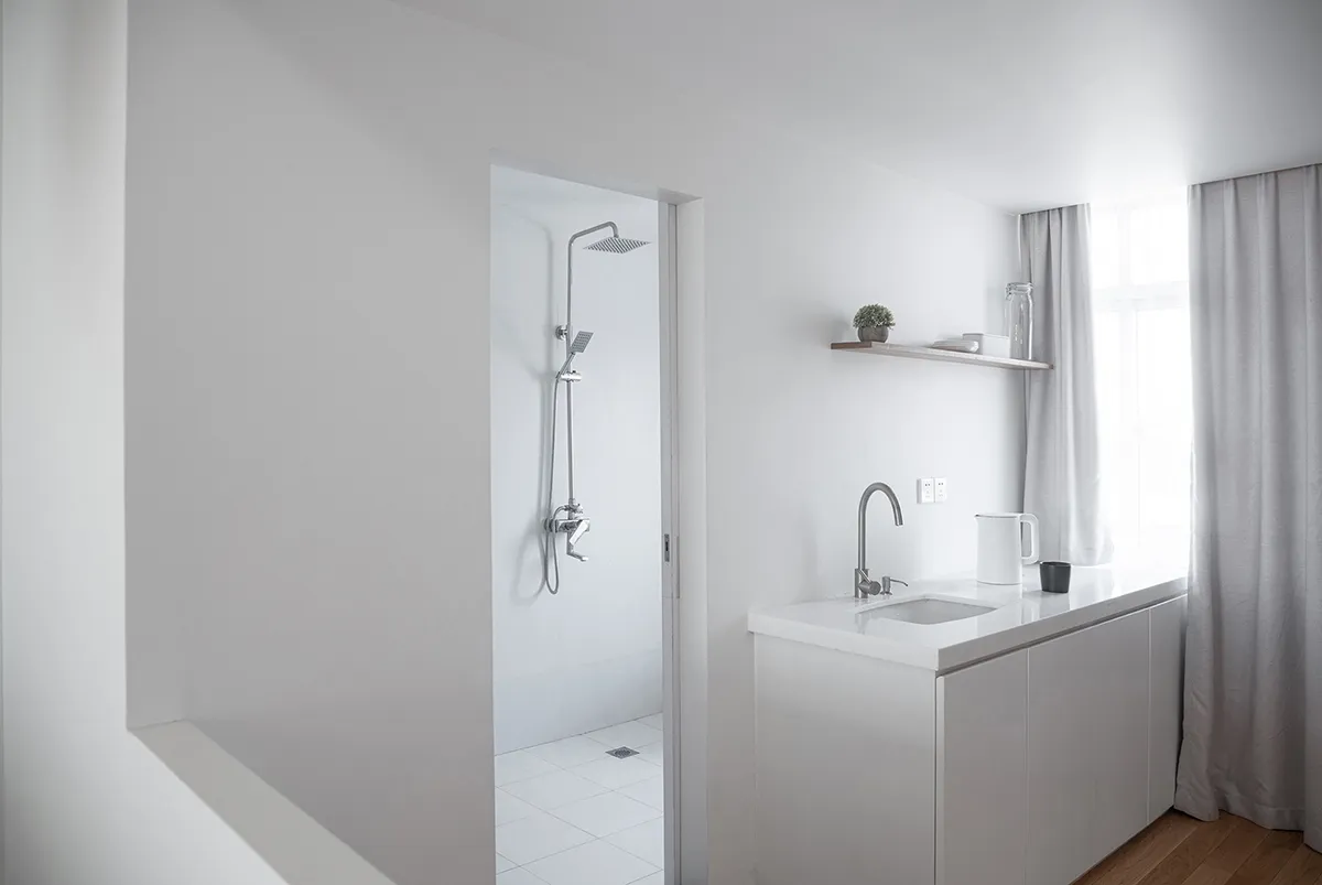 A minimalist kitchen with a white countertop, cabinets, and floor, featuring a window with sheer curtains.