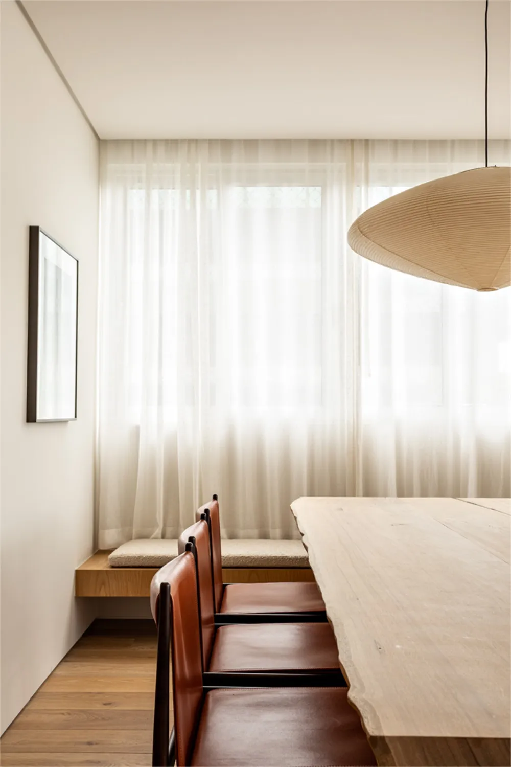 A modern minimalist dining room with a wooden table and leather chairs, featuring sheer curtains and a large pendant light.