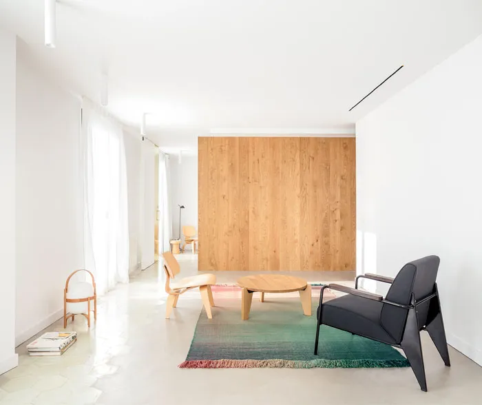 A modern minimalist living room featuring a wooden coffee table and black chair on a colorful rug.