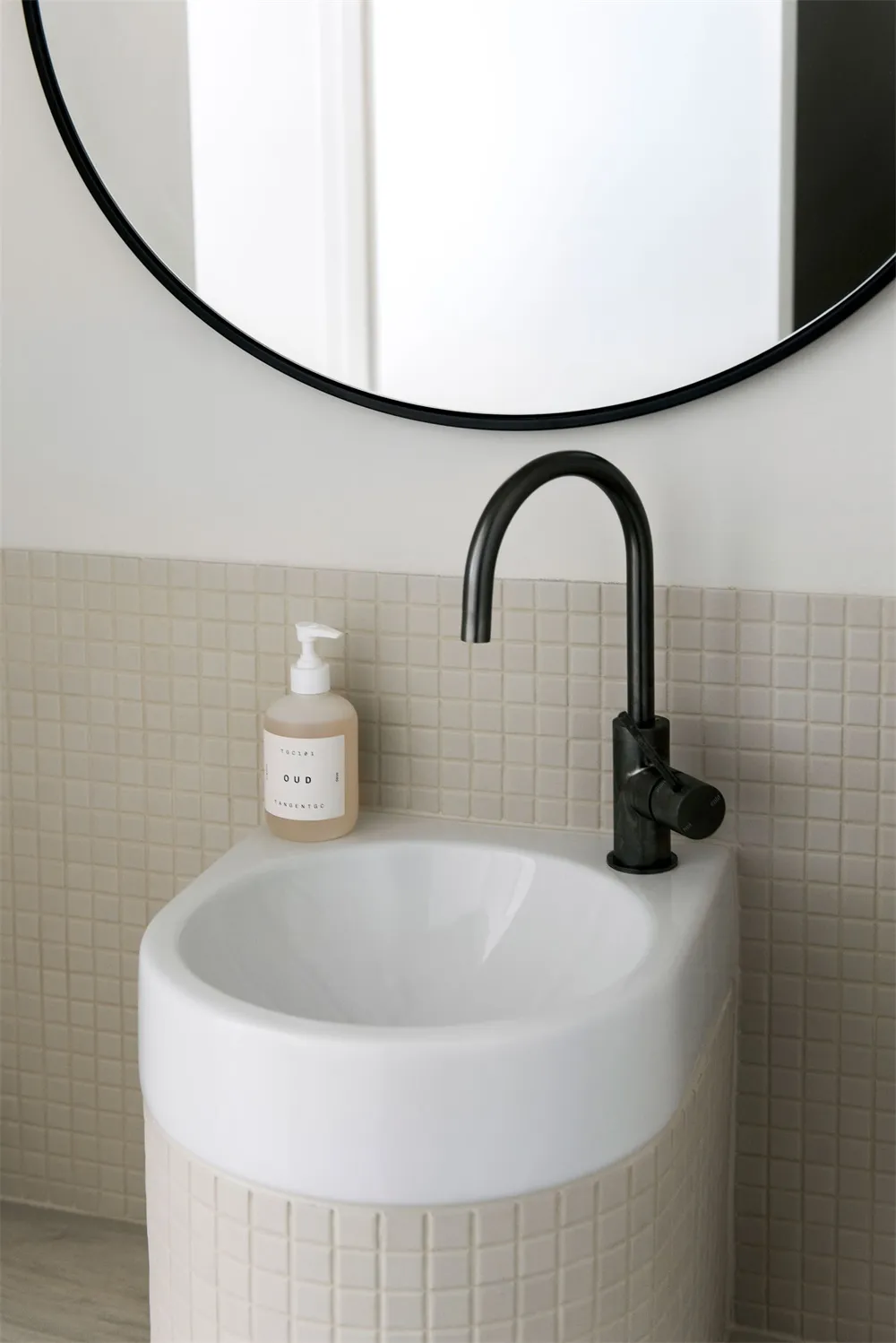 Modern minimalist bathroom featuring a sleek ceramic sink with a black faucet, beige tiled backsplash, and a round mirror abo